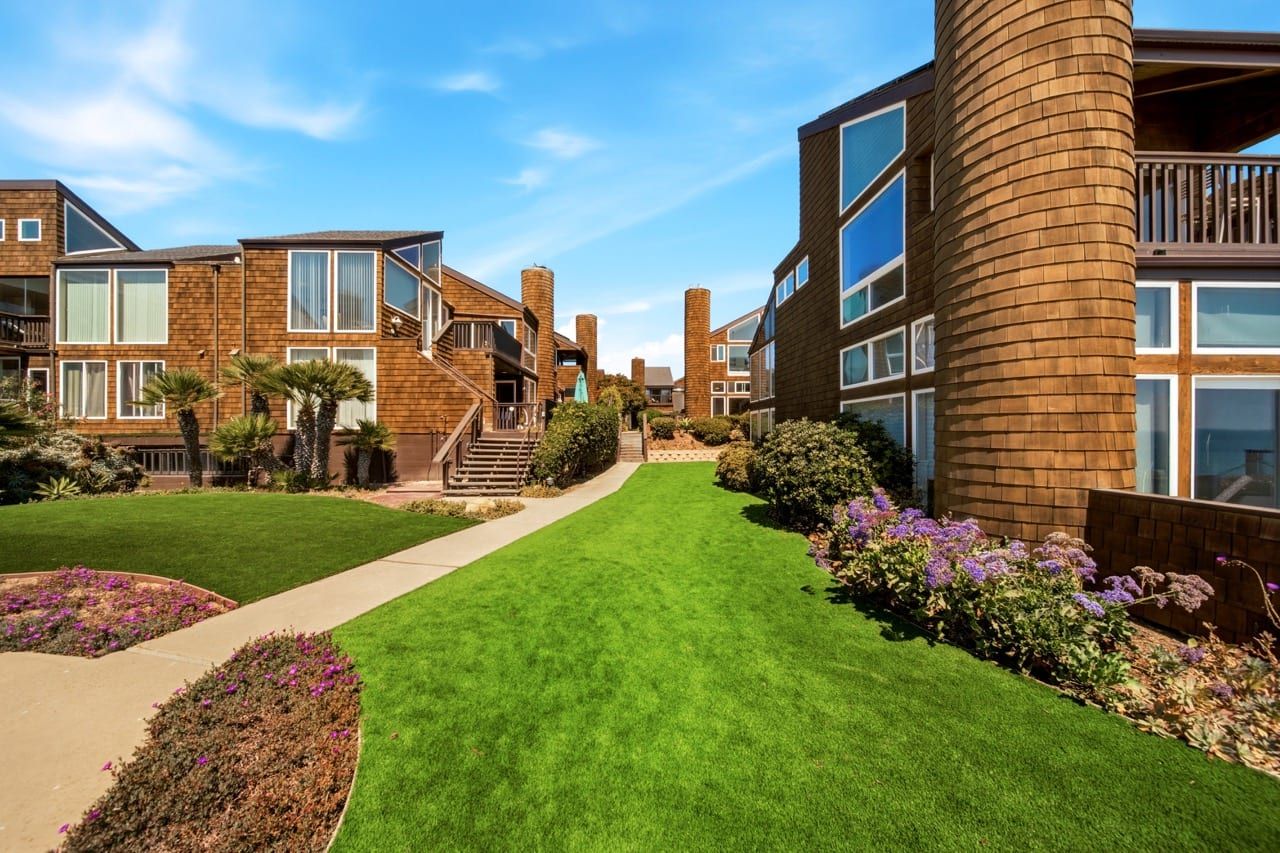 A path curving across the community lawn between the buildings, past flowering beds and palms